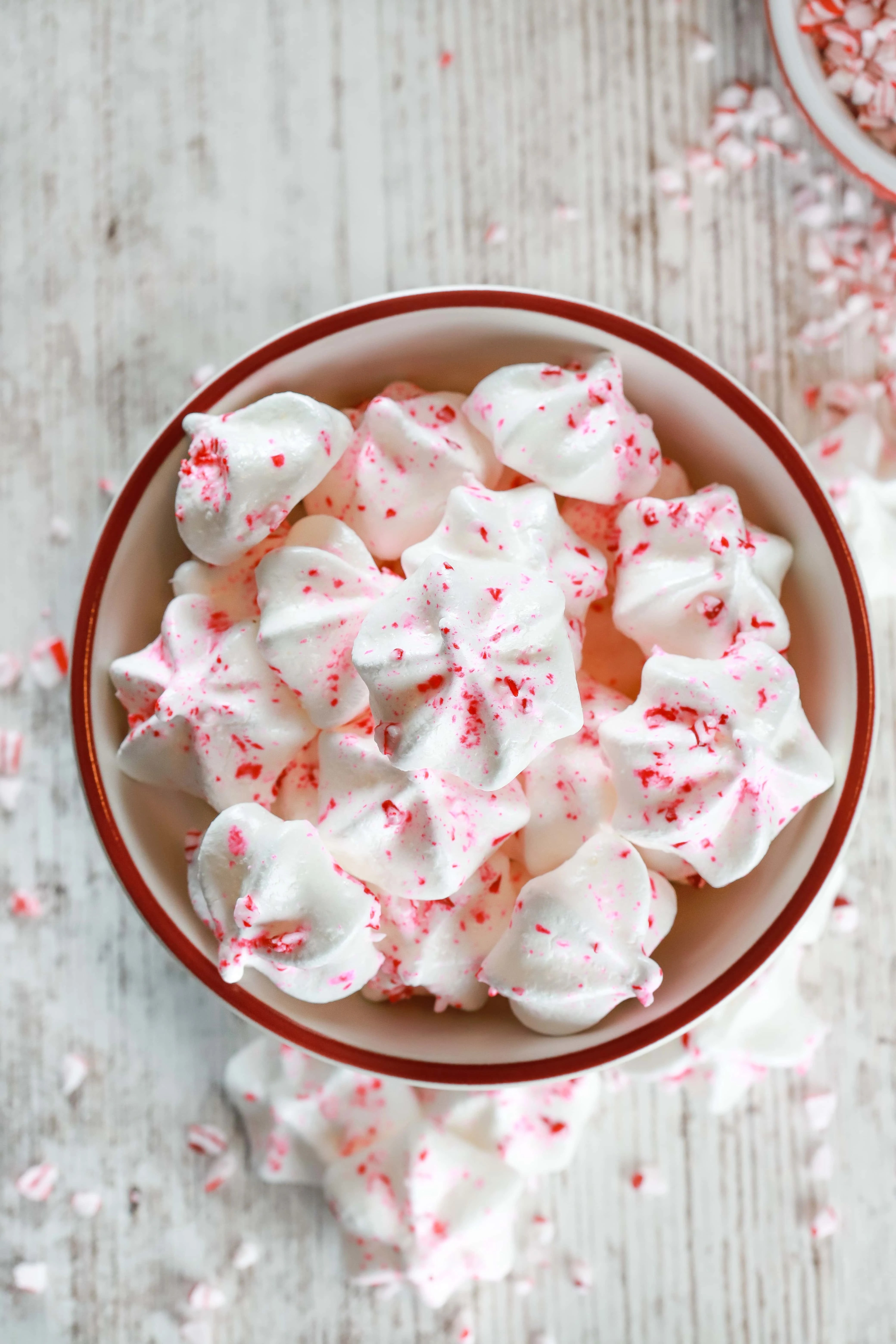 Overhead view of a white bowl of Mini Peppermint Meringue Kiss Cookies surrounded by crushed peppermint candies. Recipe for meringue cookies from A Kitchen Addiction